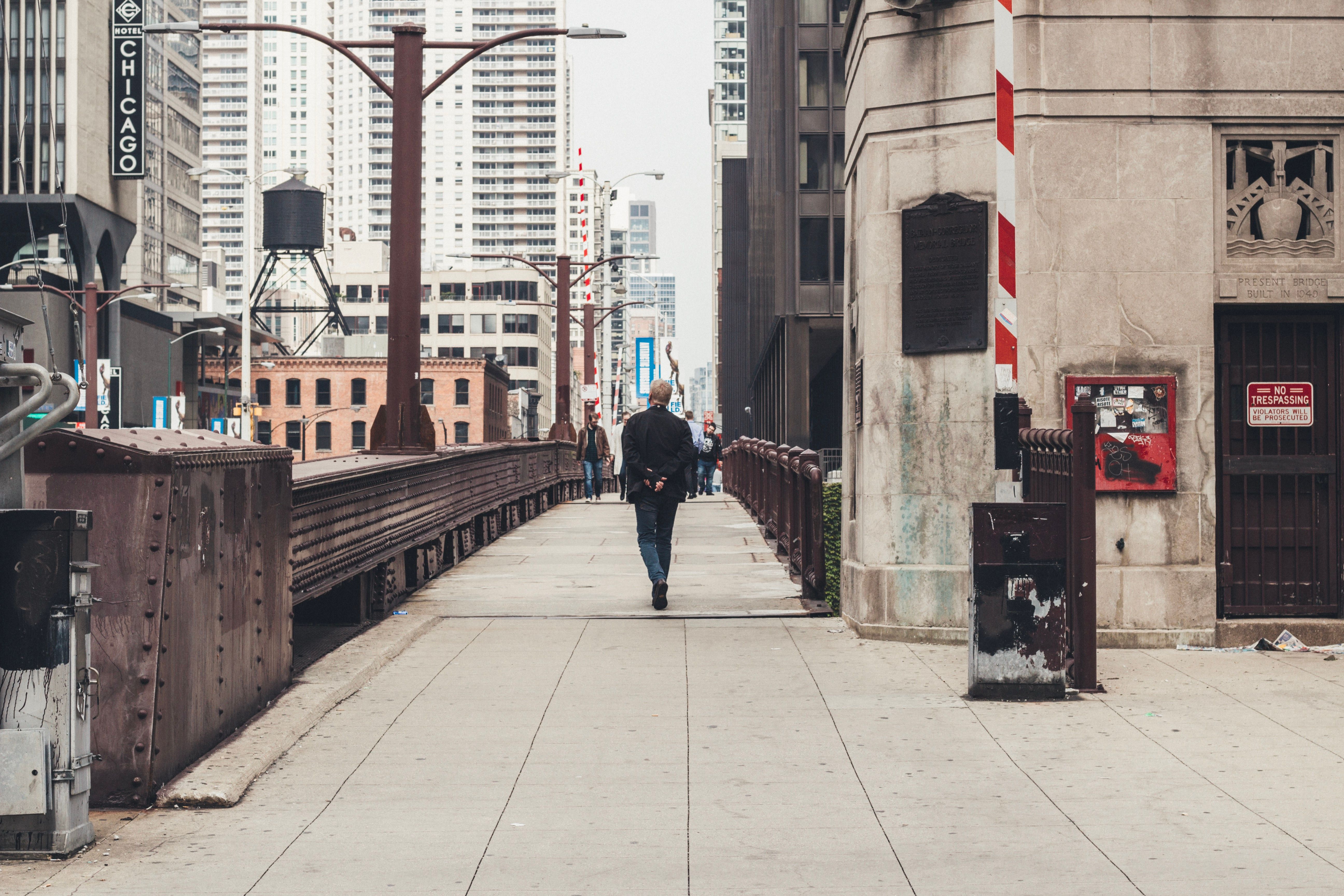 A man walking on a path adjacent to a bridge over a river in an urban setting, with tall buildings in the background.