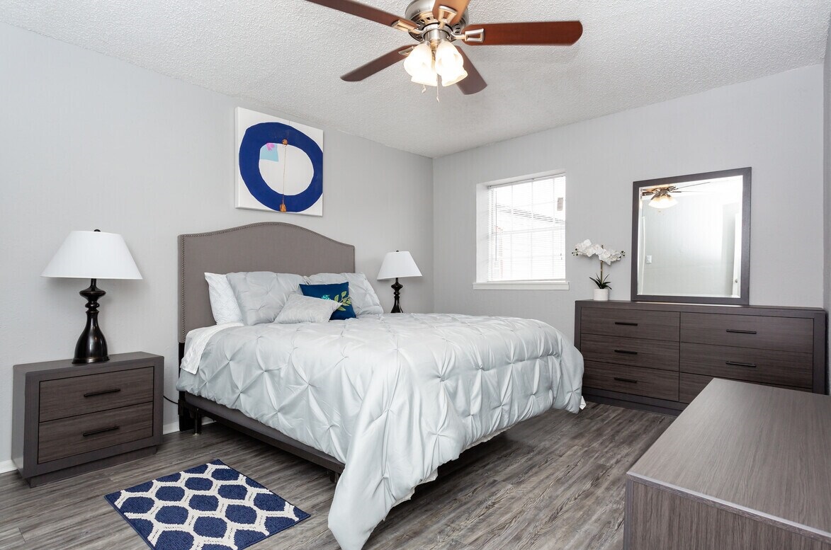 A modern bedroom featuring a gray wall, a queen-sized bed with a light comforter, matching nightstands with lamps, and a dresser. The room has wooden flooring and a blue patterned rug.
