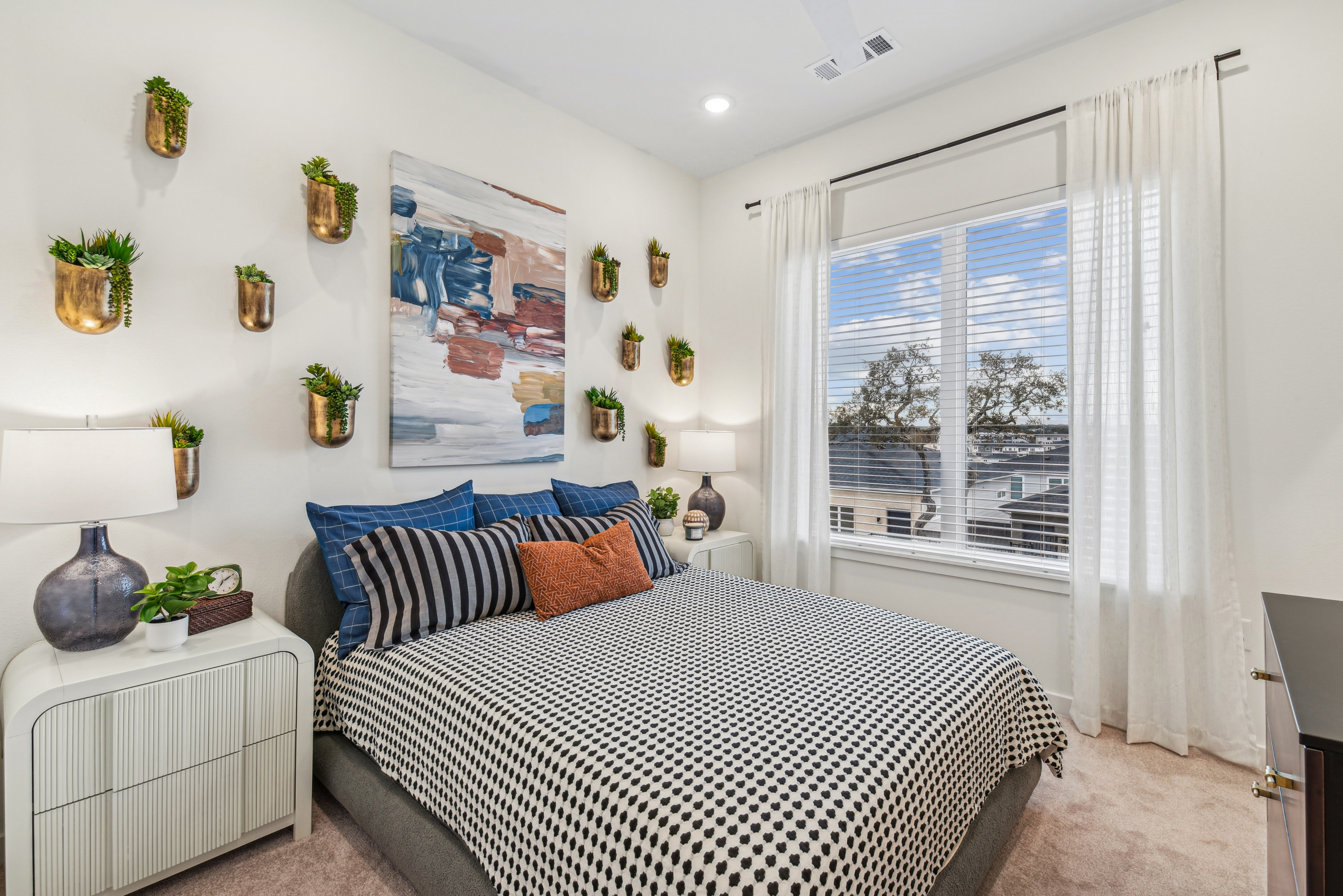 A modern bedroom featuring a bed with black and white striped bedding and decorative pillows. The walls are adorned with planters and a colorful abstract painting. Natural light enters through a window covered by sheer curtains. There's a nightstand with a lamp and a bedside plant.