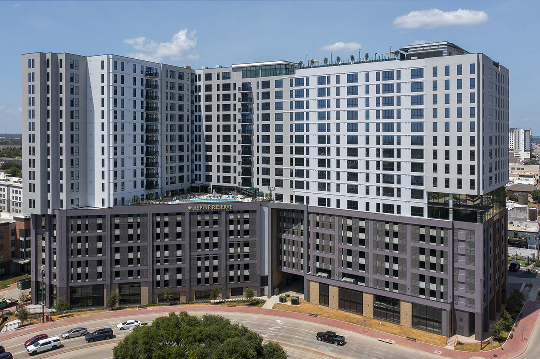 A large, modern apartment complex with multiple floors and a sleek design, featuring balconies and large windows, set against a blue sky.