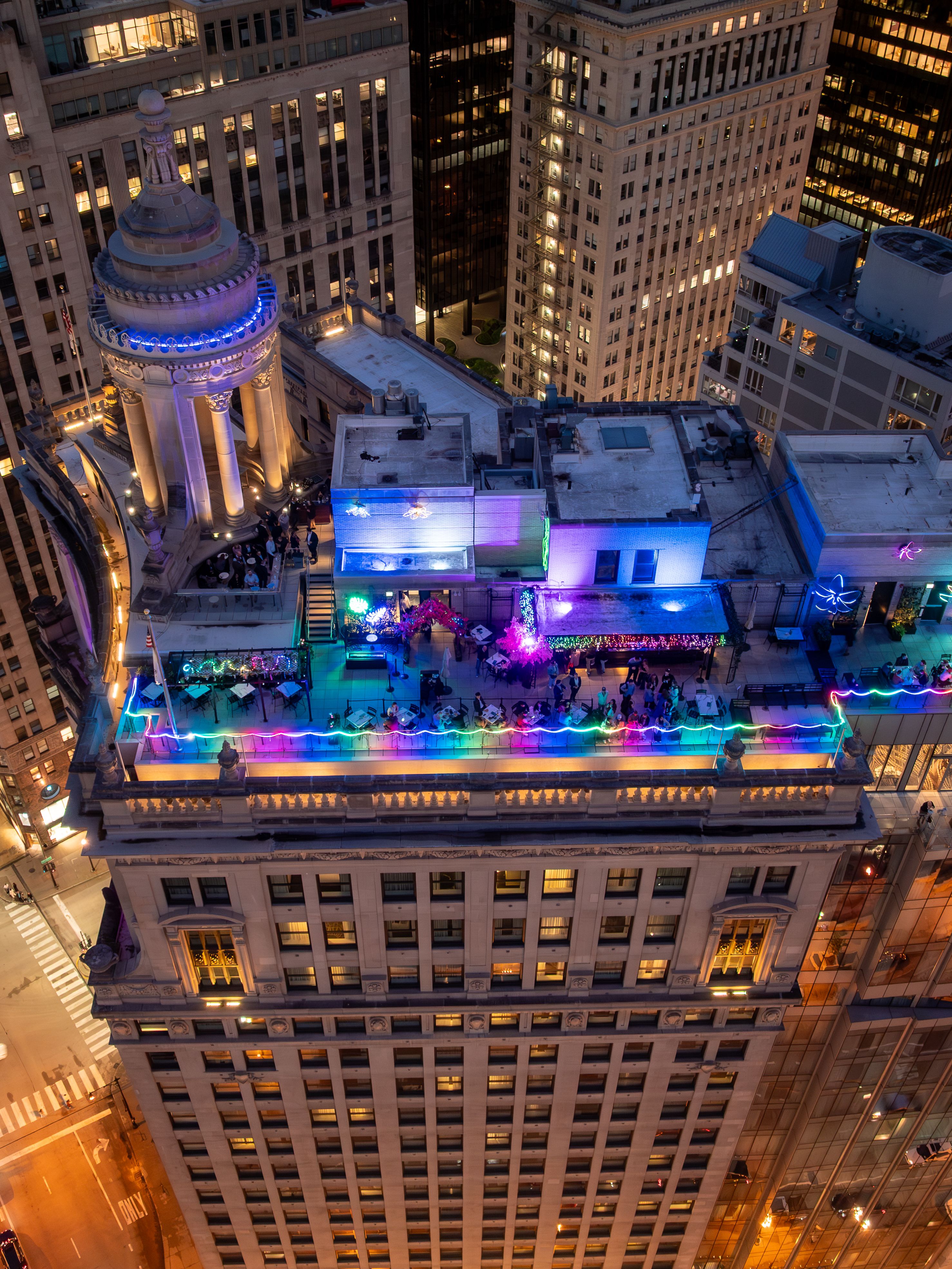 A vibrant rooftop party setting on a tall building, illuminated with colorful lights. The scene shows a bustling atmosphere with people enjoying themselves against a backdrop of city buildings at night.