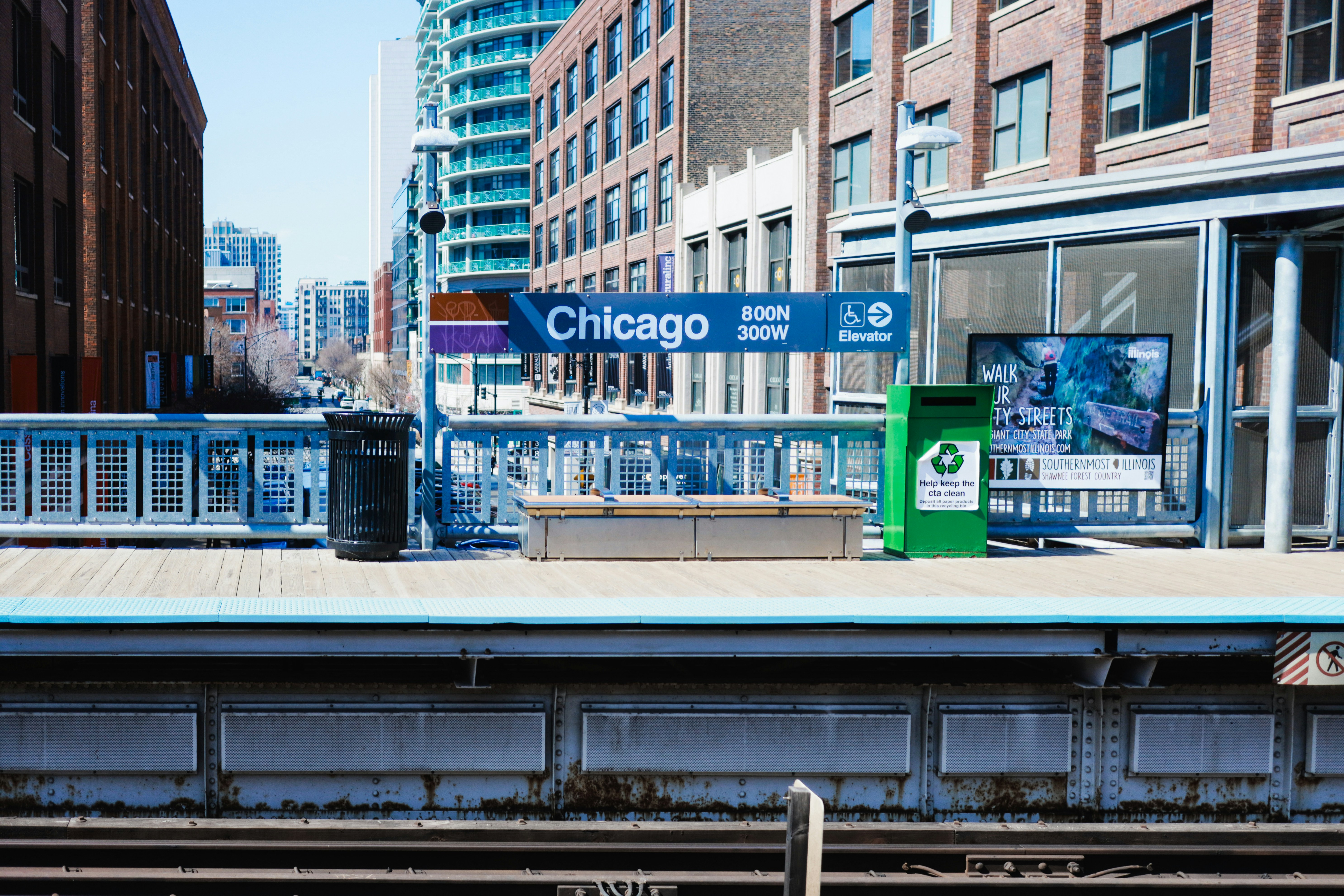 View of a train station platform with a sign indicating 'Chicago', a bench, and a trash can, surrounded by tall buildings on a clear day.