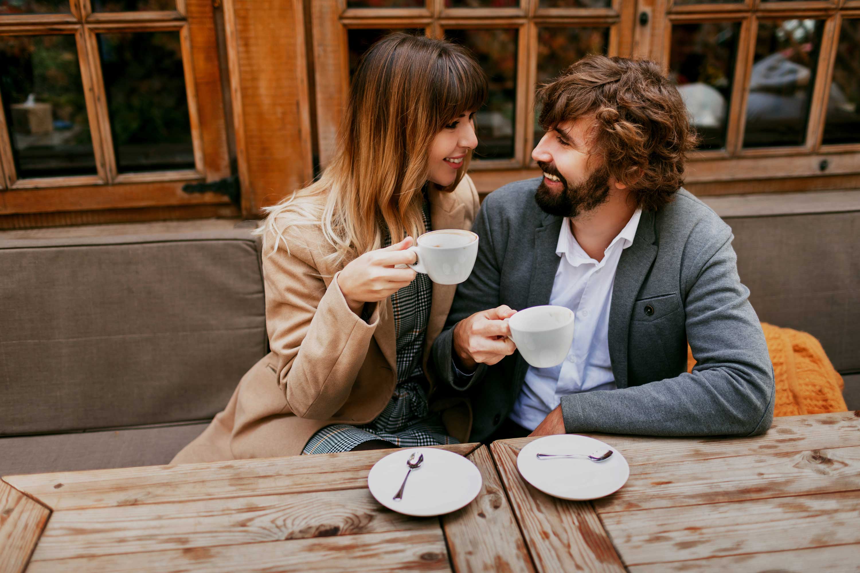A couple sharing a moment at a cozy cafe, holding cups of coffee and smiling at each other. The setting features wooden furniture and large windows.