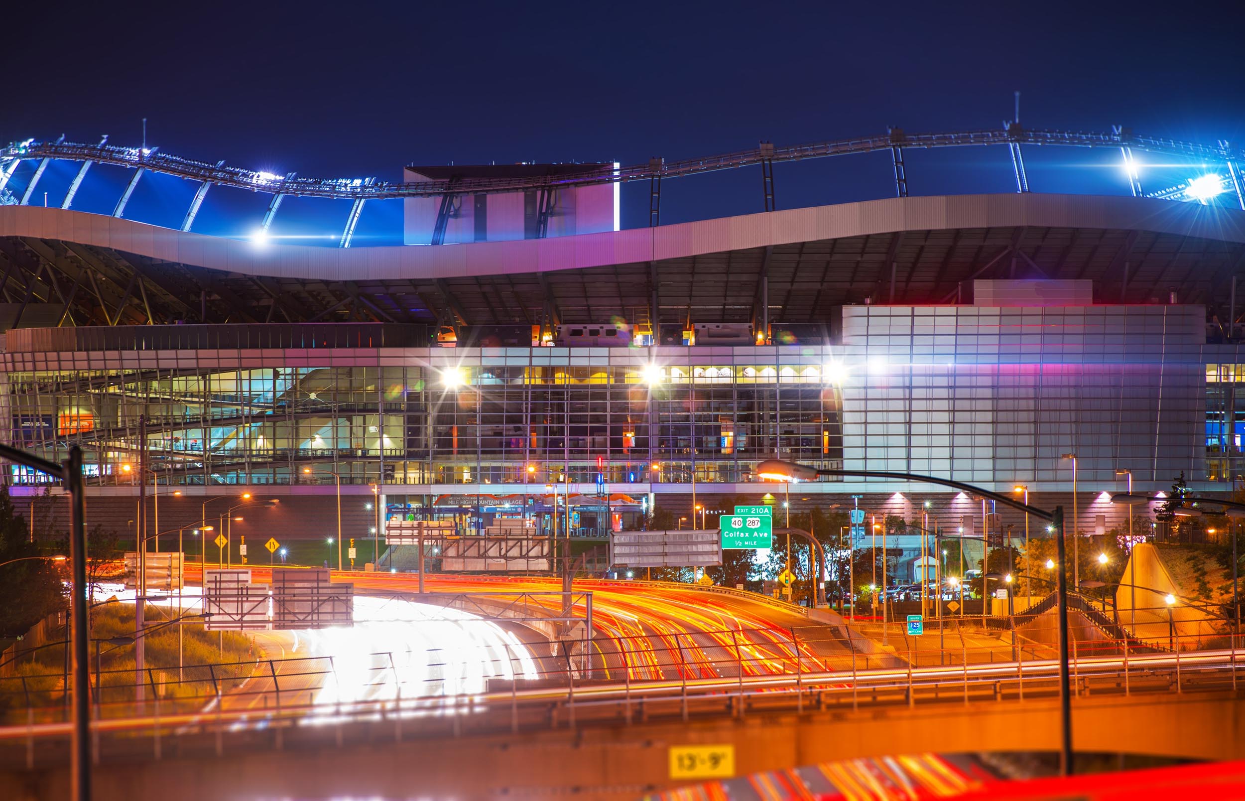 A night view of a modern stadium with bright lights and nearby highway filled with light streaks from vehicle movement.