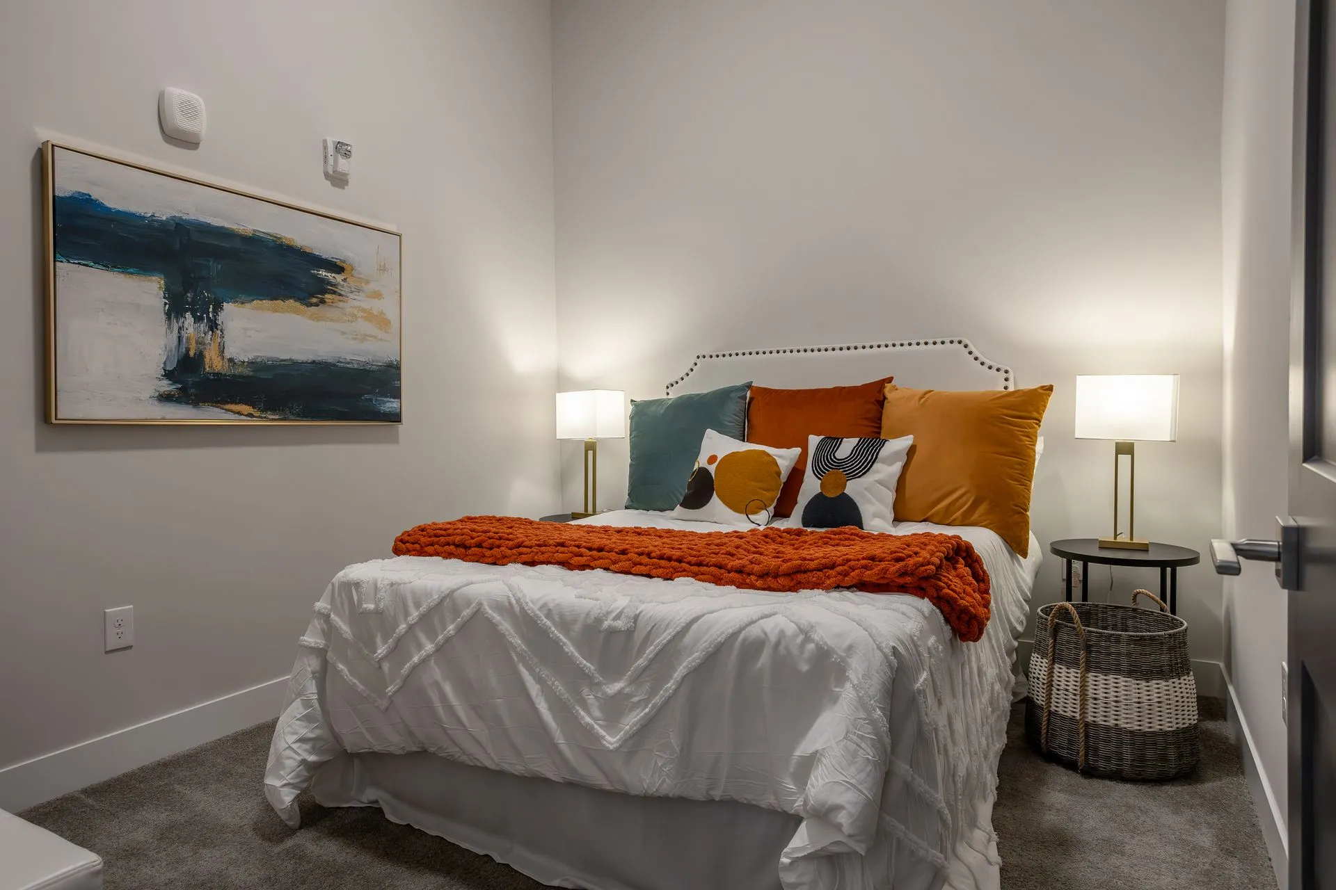 A well-decorated bedroom featuring a white bed with colorful pillows and an orange throw blanket, accompanied by two lamps on either side, and a piece of abstract art on the wall.