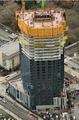 Aerial view of a tall building under construction, featuring a partially completed structure with scaffolding and safety netting on top. Surrounding roads and nearby buildings are visible.