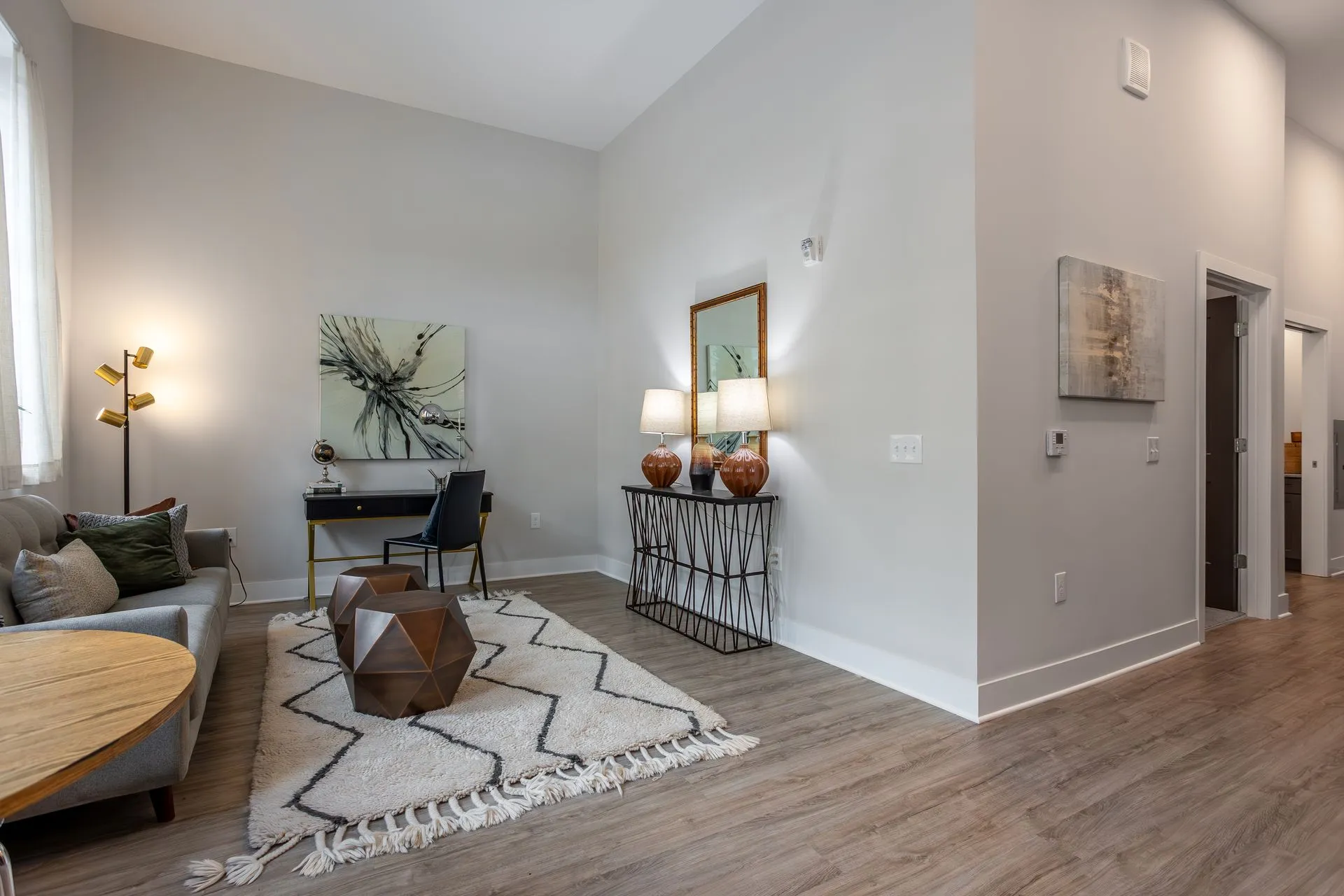 A modern living room featuring a cozy sofa, decorative lamps, a rug, and a wall art piece. A small desk is placed against the wall.