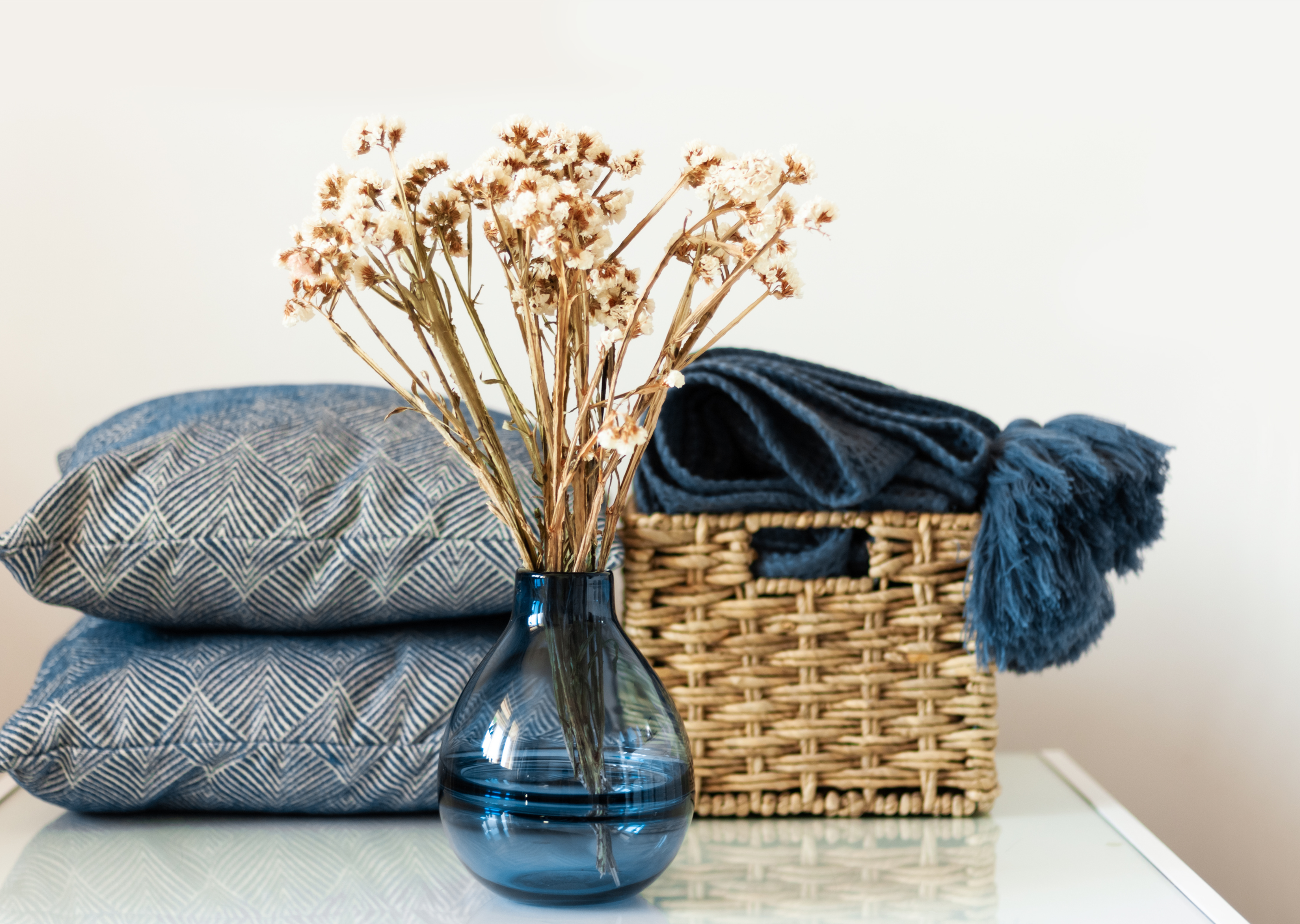 A decorative arrangement featuring a blue vase with dried flowers, two patterned blue cushions, and a woven basket filled with a soft blue throw blanket.