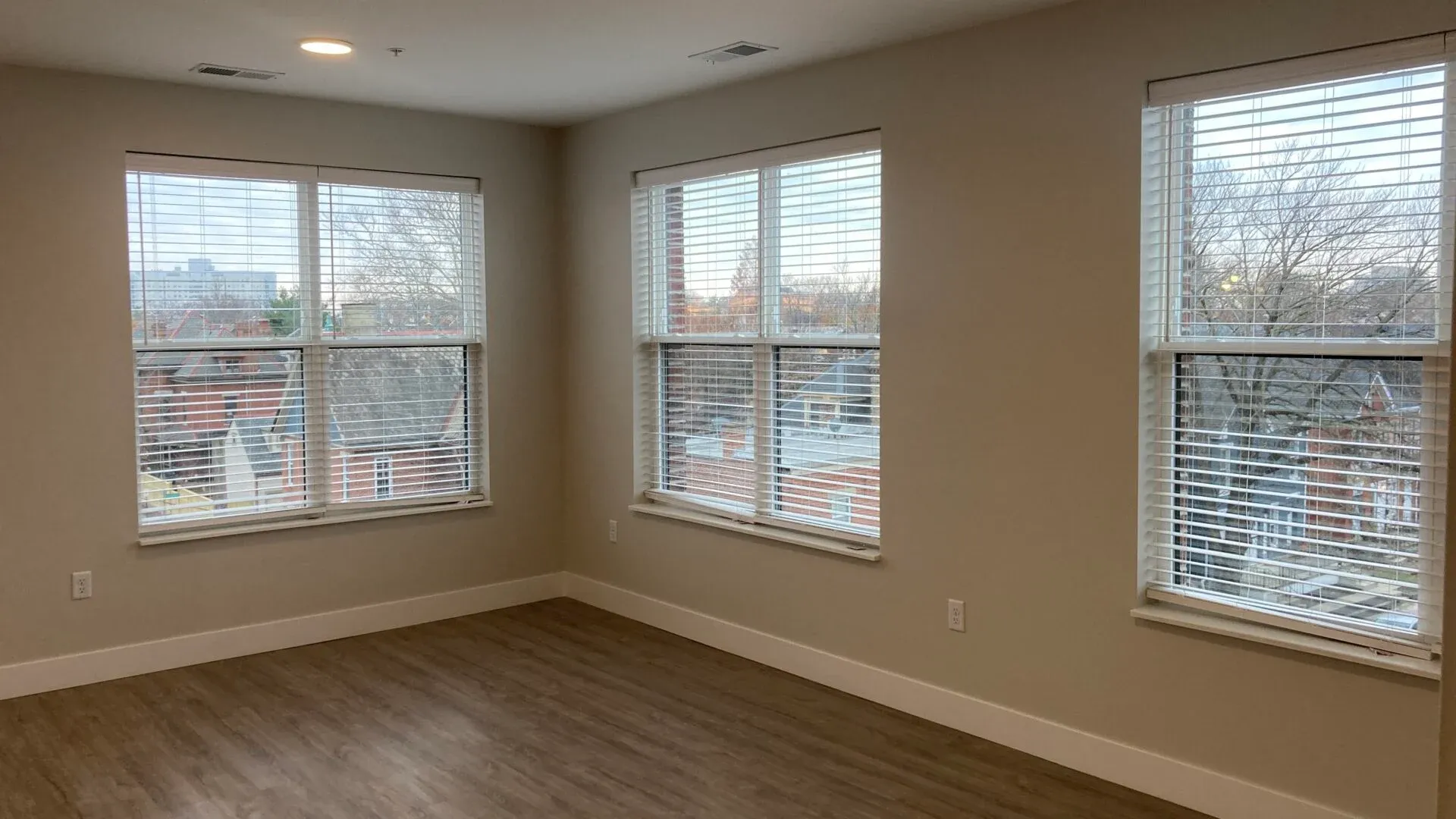 An empty room with large windows allowing natural light, featuring light-colored walls and wooden flooring.