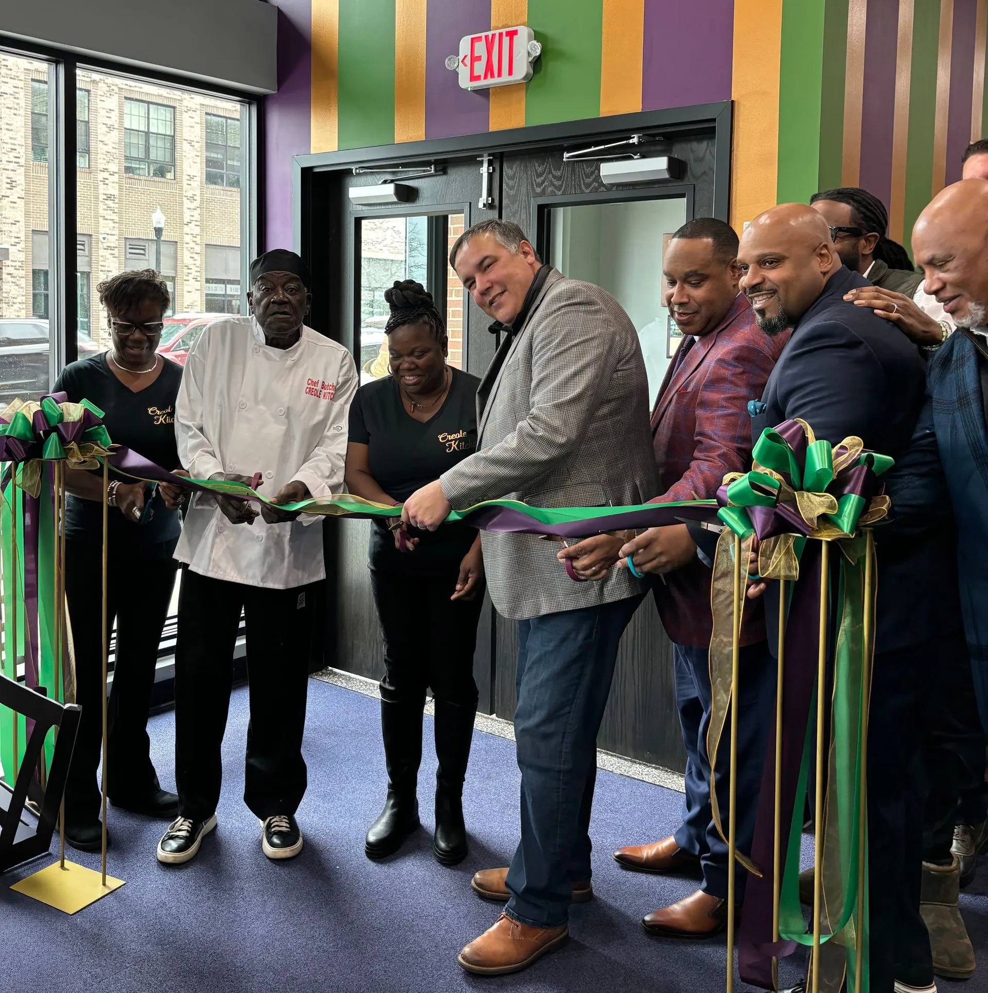 A group of people preparing to cut a ribbon in a colorful interior space. The group includes a chef in a white coat and various individuals in black shirts, displaying smiles and excitement for the event.