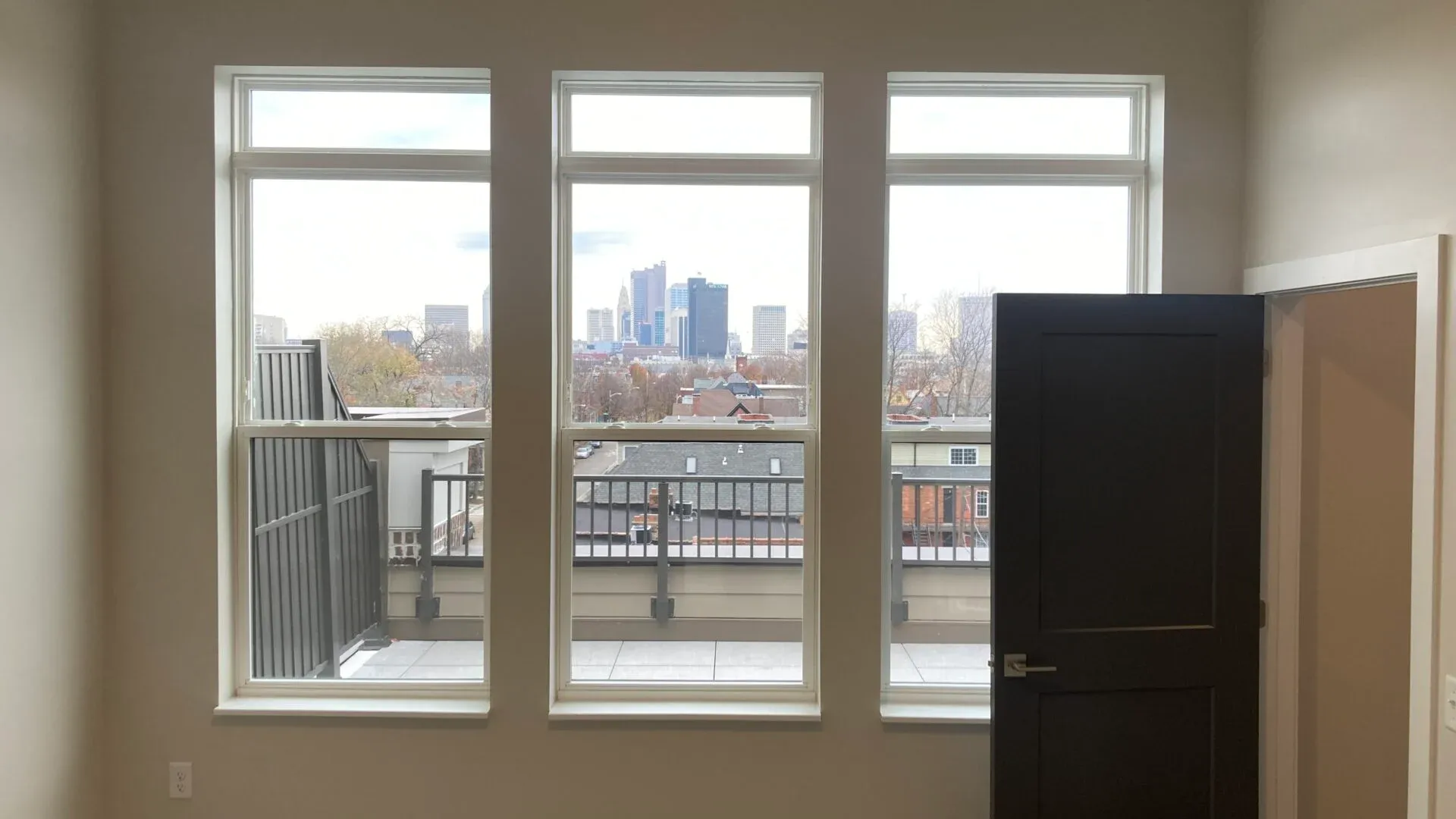 A spacious room with three large windows offering a view of a city skyline, and a door leading to a balcony.