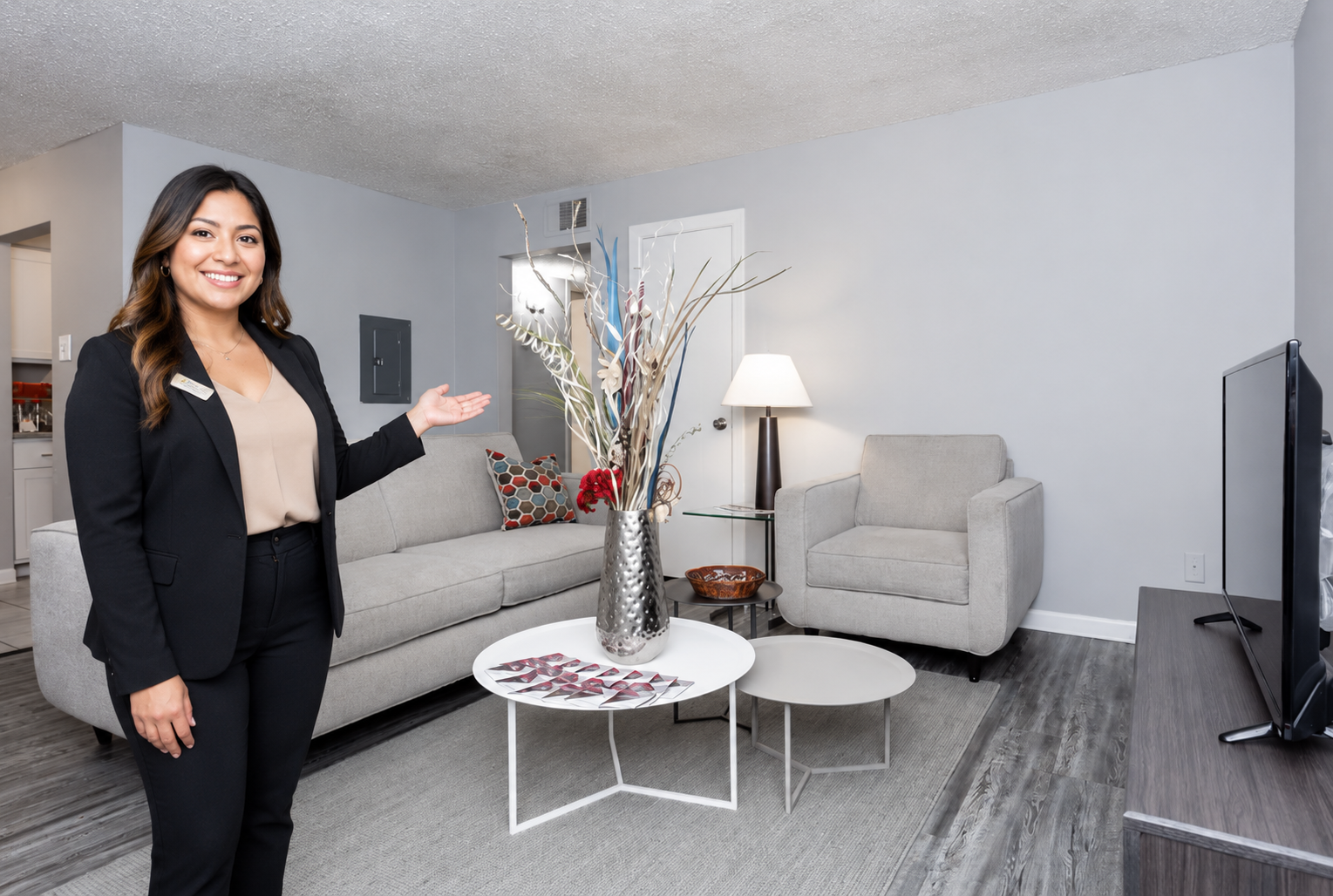 A woman in a black suit stands smiling, gesturing towards a modern living room with light gray walls, a gray sofa, and a coffee table with decorative elements. A floor lamp and a television are also visible.