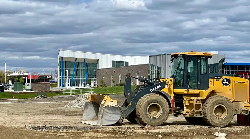 A construction site with a yellow bulldozer in the foreground and a modern building in the background under cloudy skies.