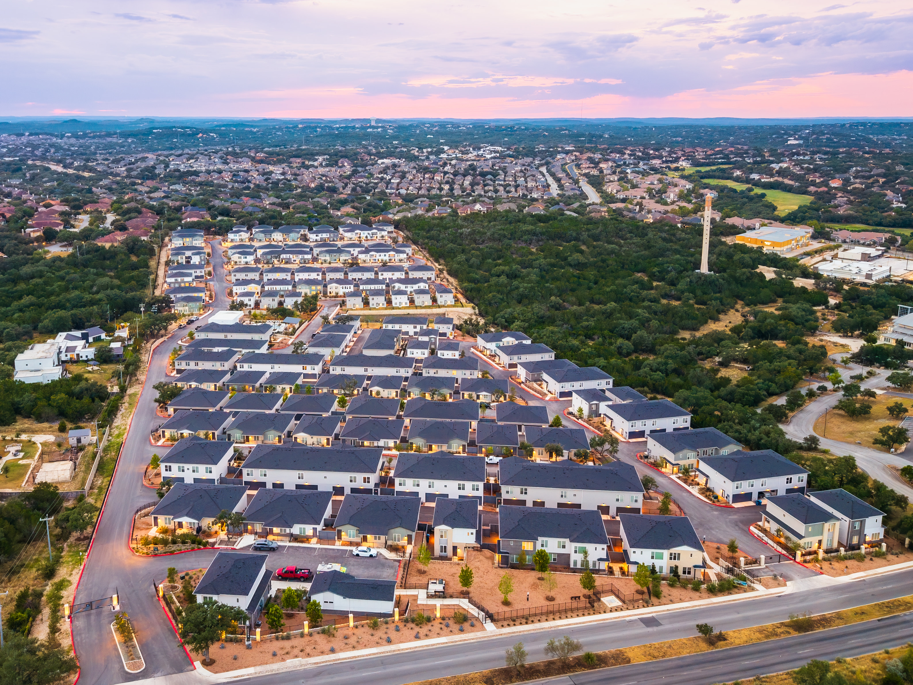 Aerial view of a residential area featuring multiple houses, trees, and roads, with a distant view of a cityscape and a tower.