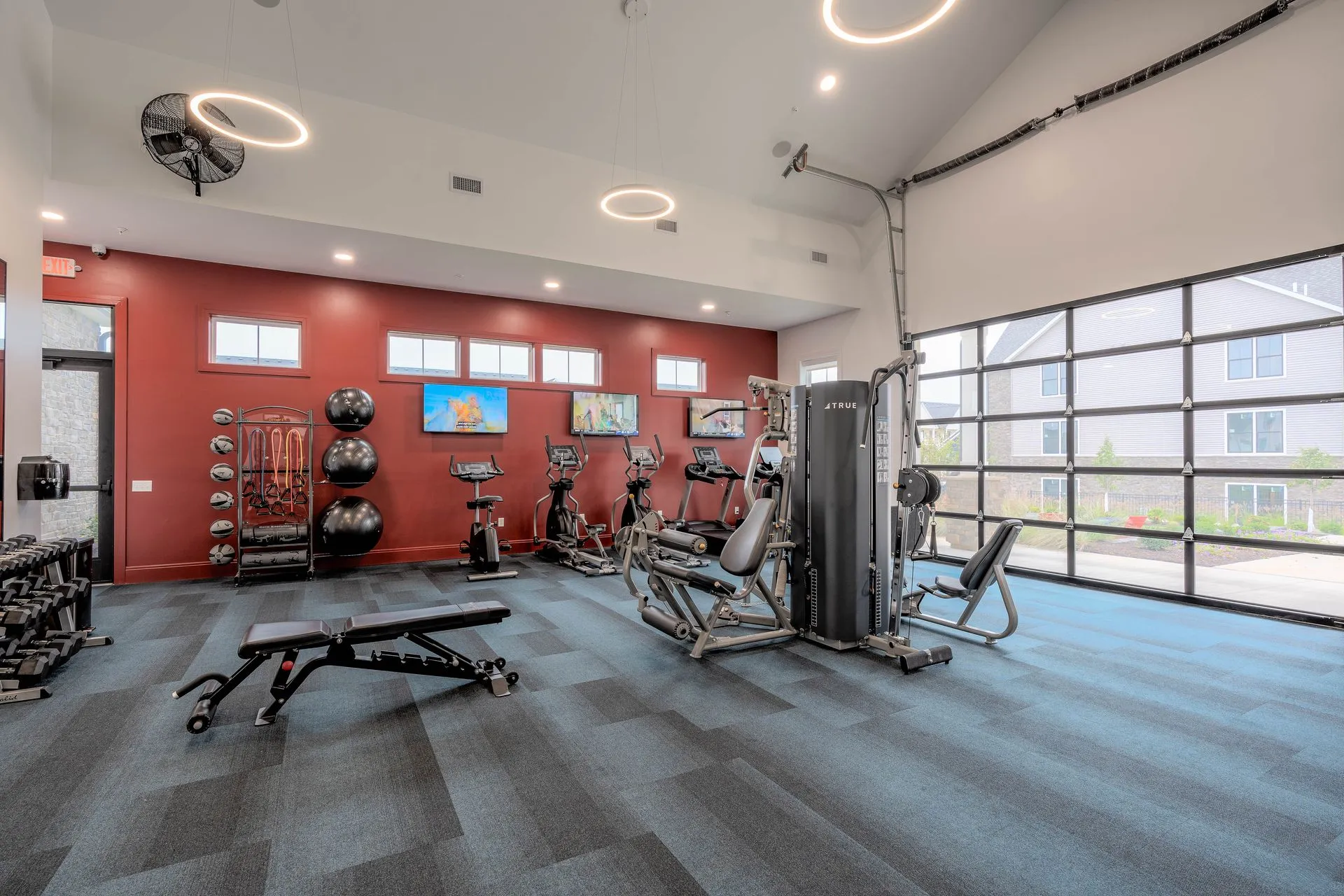 A modern gym space with exercise equipment including weights, cardio machines, and a multi-gym setup. The décor is minimalistic with neutral colors and large windows allowing natural light.