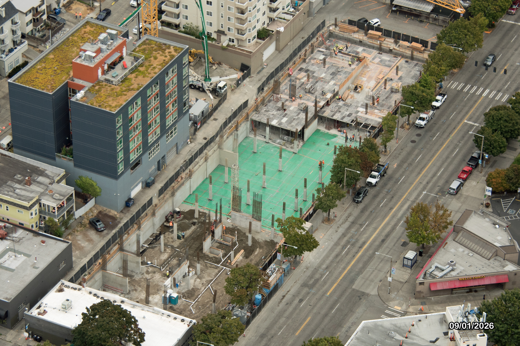 Aerial view of a construction site with visible foundations, surrounded by existing buildings and a busy street.