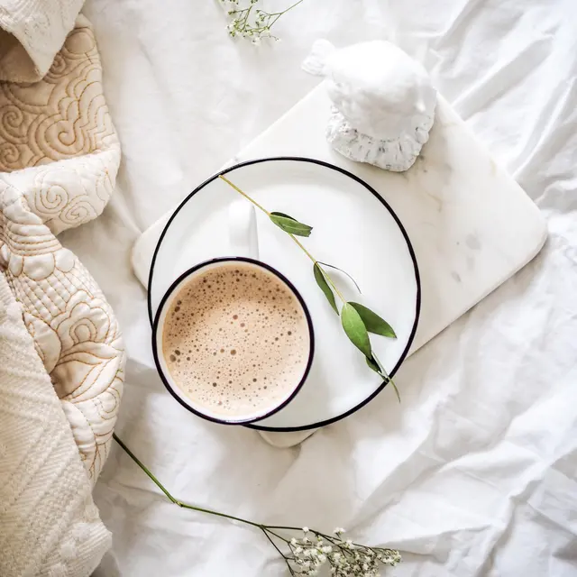 A cozy setup featuring a cup of coffee on a marble tray, surrounded by greenery and soft fabrics.