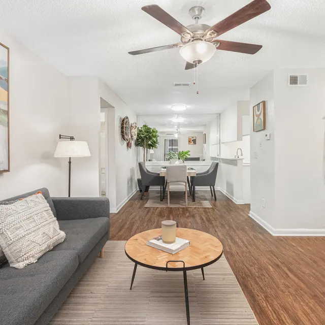 A modern, well-lit living room with a ceiling fan, artwork, a grey couch, decorative pillows, a coffee table, and a dining area in the background.