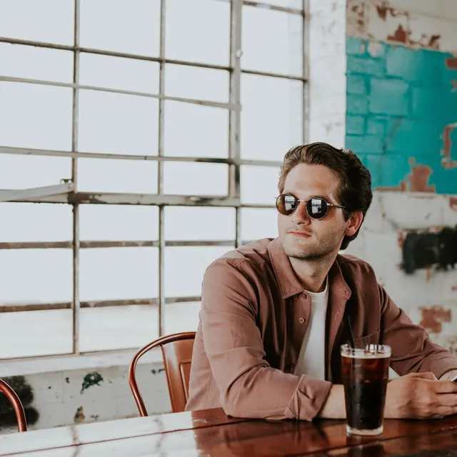 Vernon Court A man wearing sunglasses sits at a wooden table in a spacious, industrial-style café, holding a phone and a drink.
