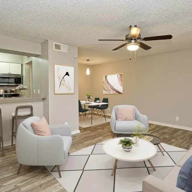 Modern living room with light gray armchairs, a round white coffee table, wooden flooring, and an open kitchen in the background.
