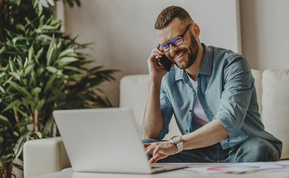 Man sitting on a couch, smiling while talking on a phone and working on a laptop, surrounded by houseplants.