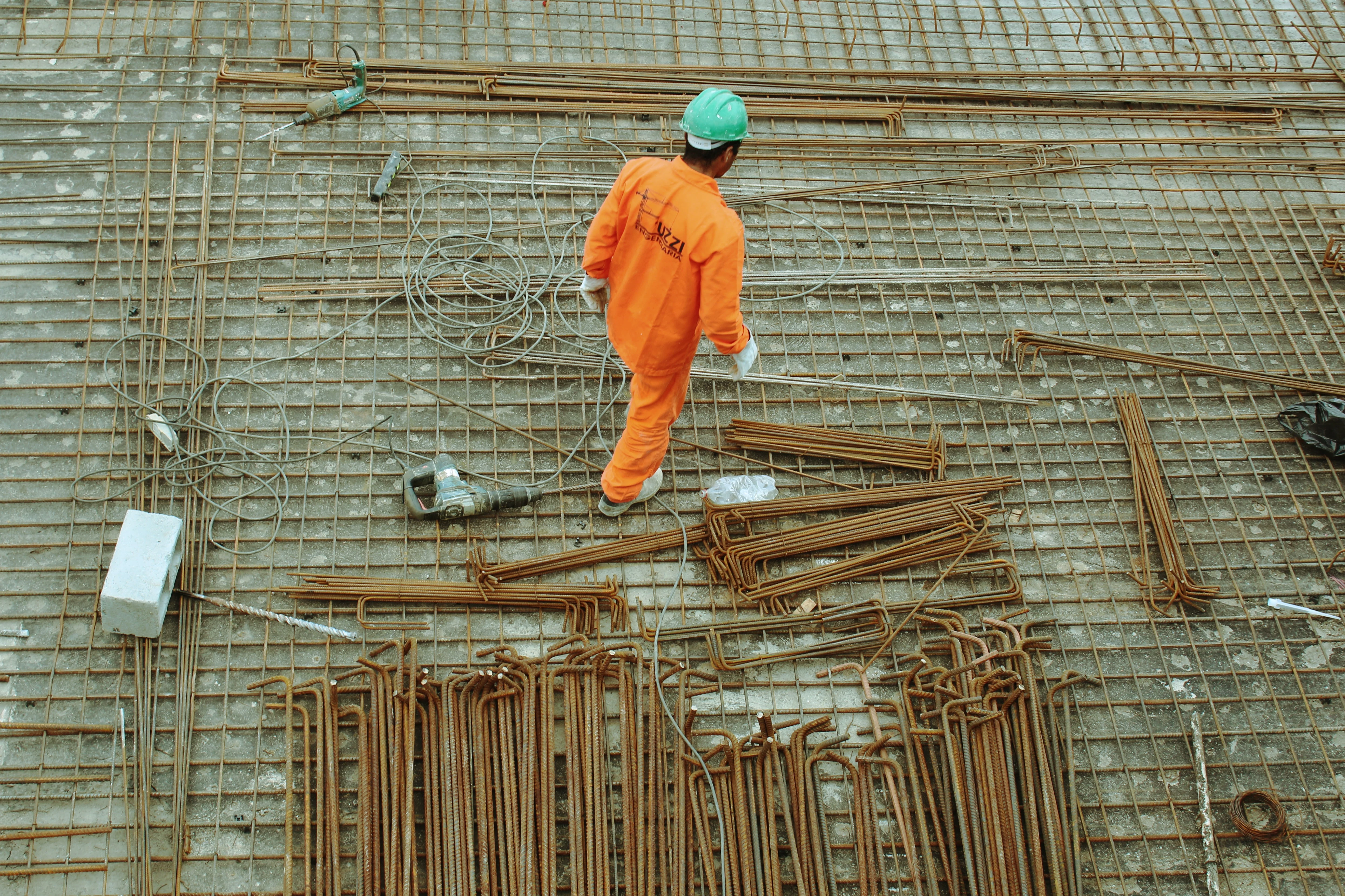 A construction worker in an orange jumpsuit and green helmet walking on a steel rebar grid. Tools and materials scattered around.