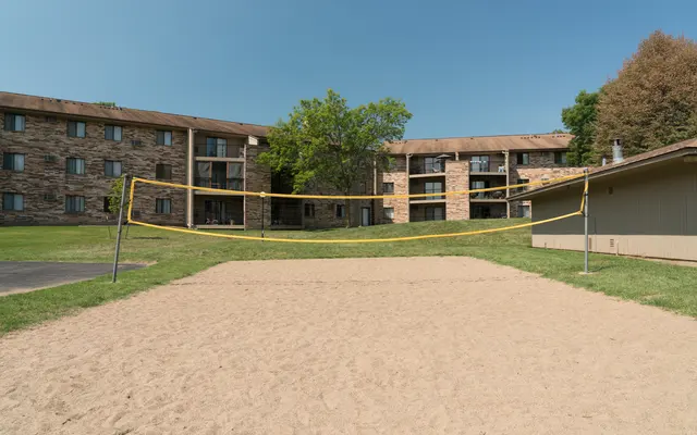 an exterior view of a Volley Ball Court with the sand ground, green grass on the sides, trees, and a view of the apartment buildings - Grass, Plant