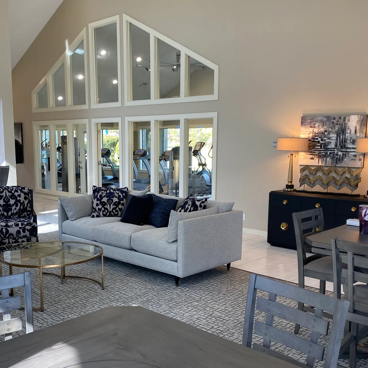 The Hunt A modern communal lounge with sofas, tables, a bar area, and large windows revealing an adjacent fitness room with exercise equipment.