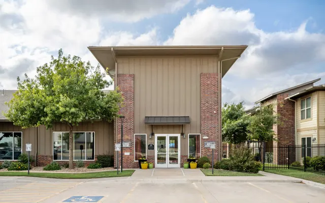 Main entrance of a modern building with a brick facade and large windows, surrounded by trees and landscaped areas.