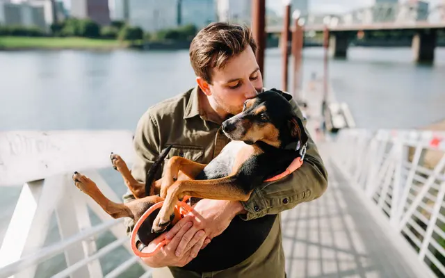 Man in a green shirt lovingly holds and kisses a small black and brown dog on a riverside walkway with a city skyline in the background.