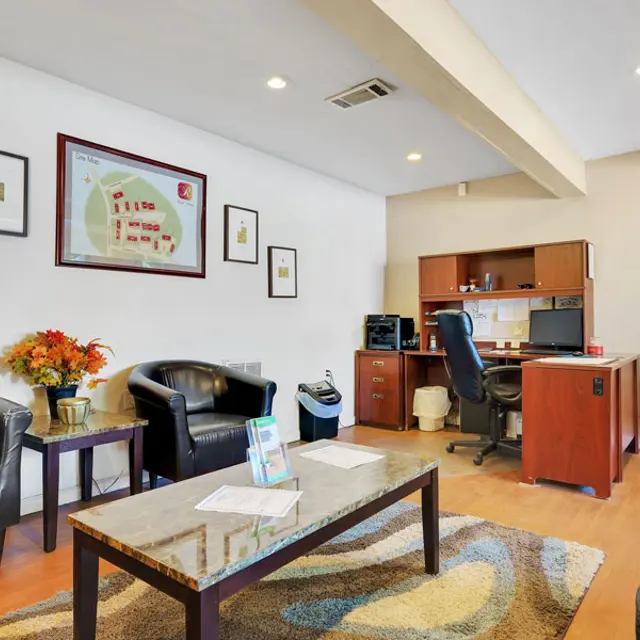 A modern office lobby with two black leather chairs, a wooden coffee table, and a desk area containing a computer and office supplies. The walls are decorated with framed pictures and a map. There is a small plant on the side table.