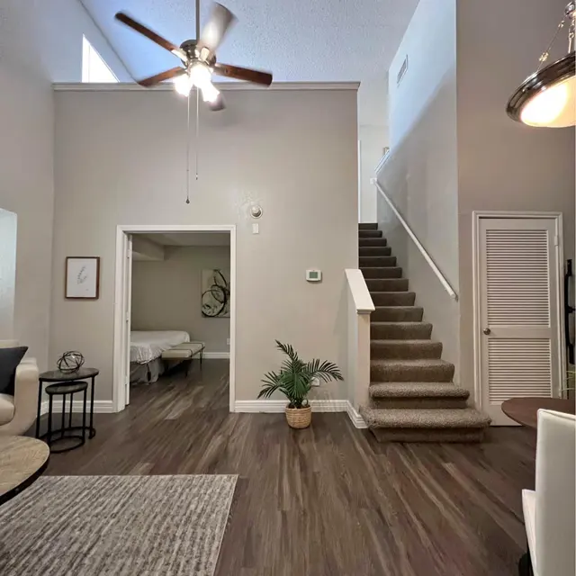 An interior living space featuring a ceiling fan, a staircase, and a small plant. The floor has dark wooden planks and a large area rug, with a doorway leading to another room and white walls.