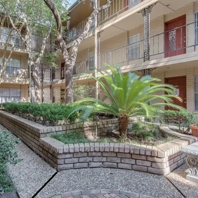 A courtyard area in an apartment complex featuring a palm tree surrounded by greenery and brick pathways.