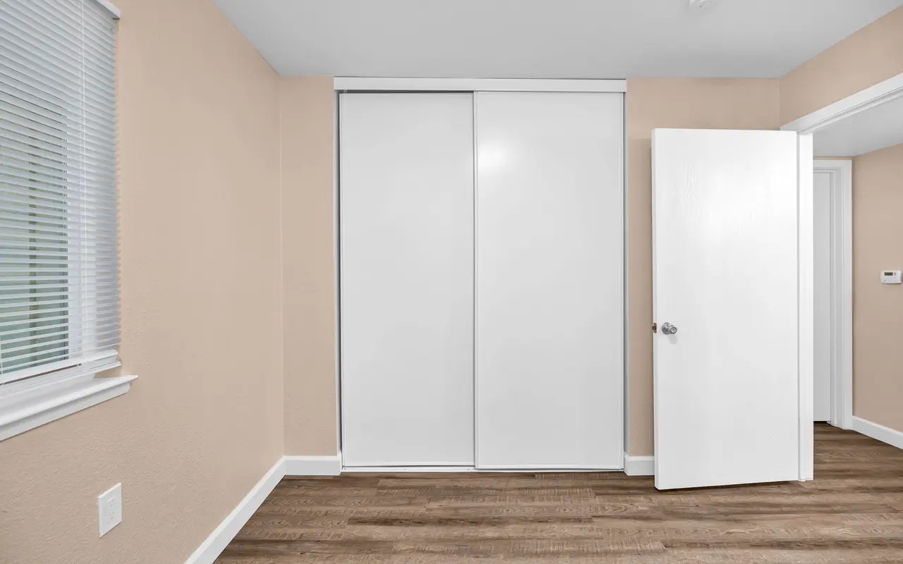 Minimalistic interior view of a room with light brown walls, wooden flooring, sliding closet doors, and an open door to another room.