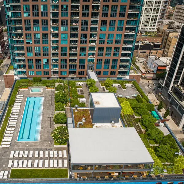 Aerial view of a modern rooftop with a swimming pool, gardens, and lounge chairs, surrounded by tall city buildings.
