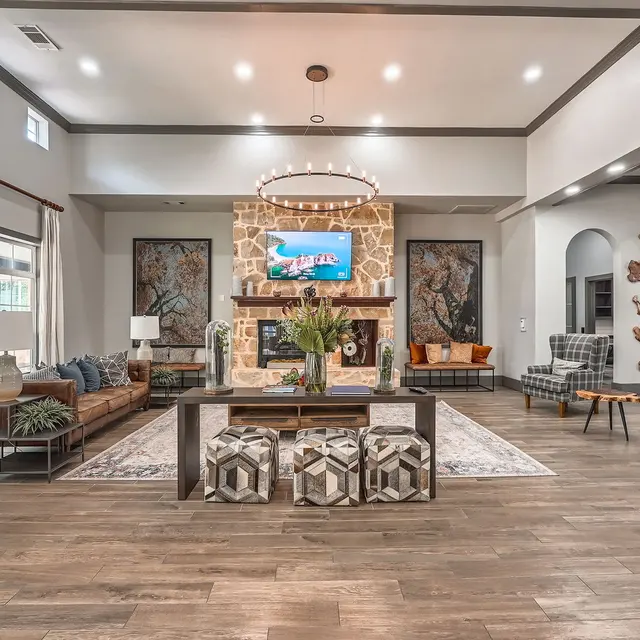 A spacious and stylish living room featuring a large brown sofa, decorative chairs, and a coffee table with geometric patterned ottomans. The room has bright décor, a large TV above a stone fireplace, and art on the walls, creating a warm and inviting atmosphere.