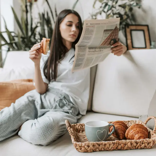 A woman sits on a couch reading a newspaper, enjoying a pastry, with a cozy setting and a tray of croissants nearby.
