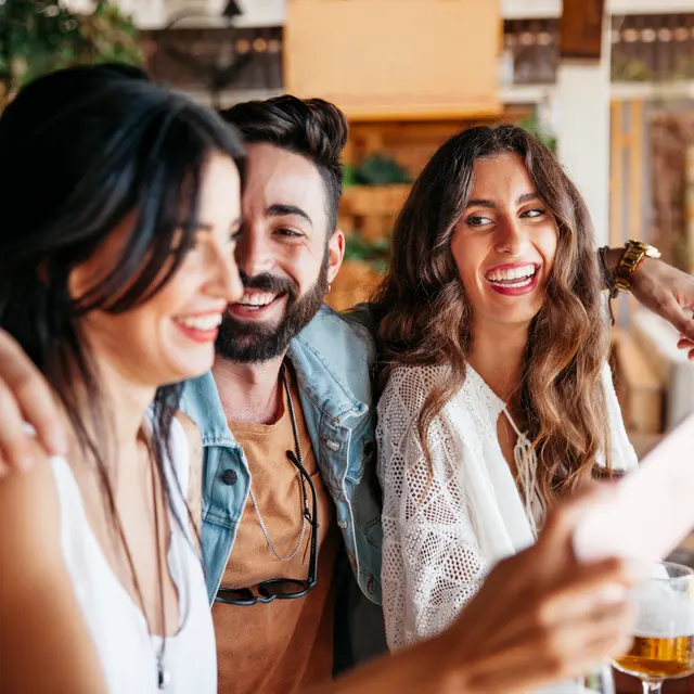Three friends laughing and taking a selfie at a bar, with drinks in front of them.