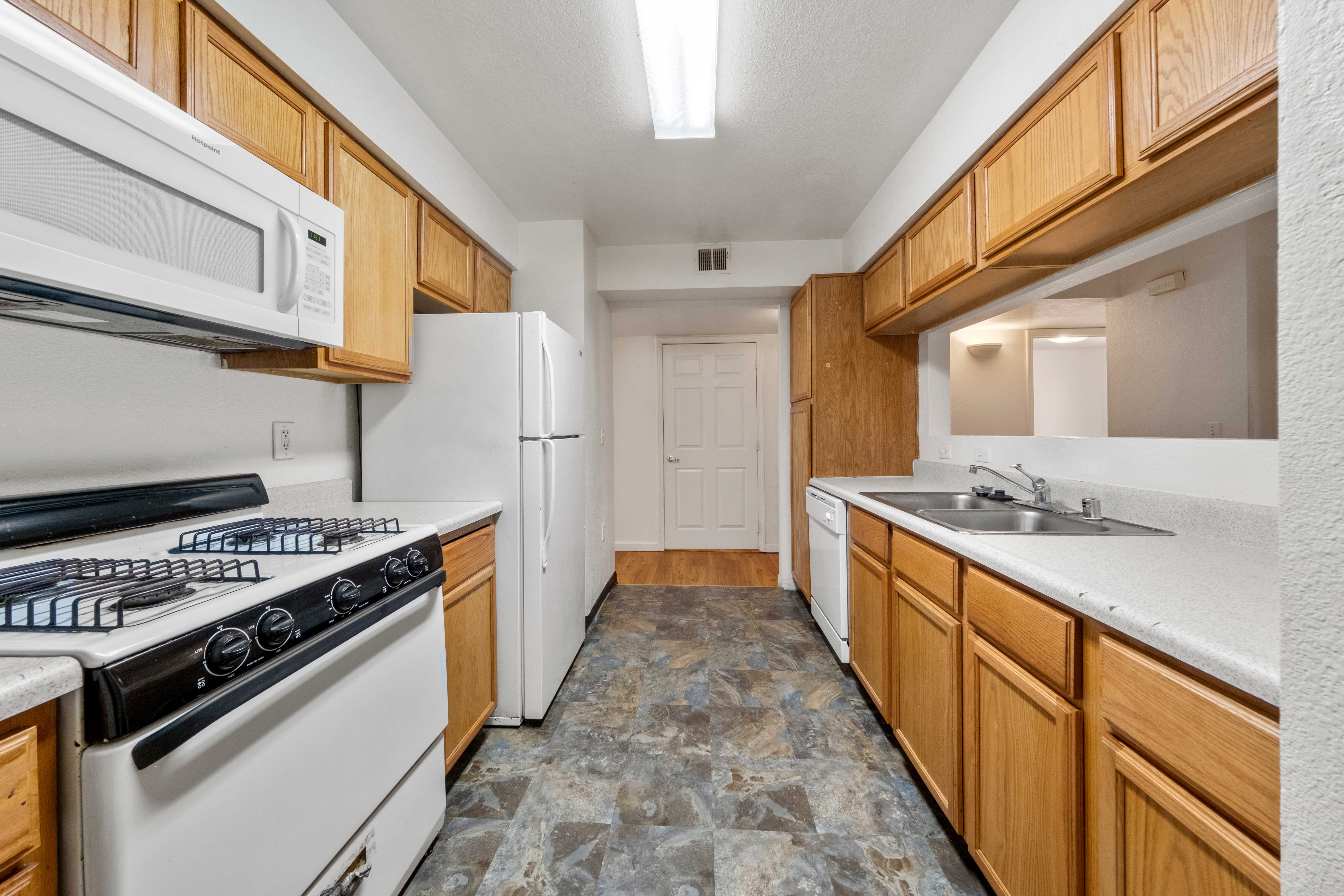 Sheridan Apartments Kitchen A galley-style kitchen featuring wood cabinetry, a double sink, and essential appliances, including a gas stove, microwave, refrigerator, and dishwasher. The kitchen has ample countertop space and a functional layout with a tile floor, creating an efficient and practical cooking area.