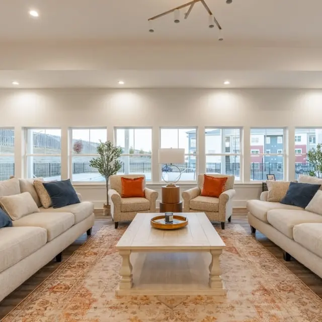 A modern living room with two light-colored sofas facing each other. The room features large windows providing natural light and a view outside. A light wooden coffee table is centered between the sofas, accompanied by decorative items. Two small plants are positioned in the corners, and decorative cushions in orange and dark blue colors complement the sofas. Soft lighting illuminates the space, enhancing its inviting atmosphere.