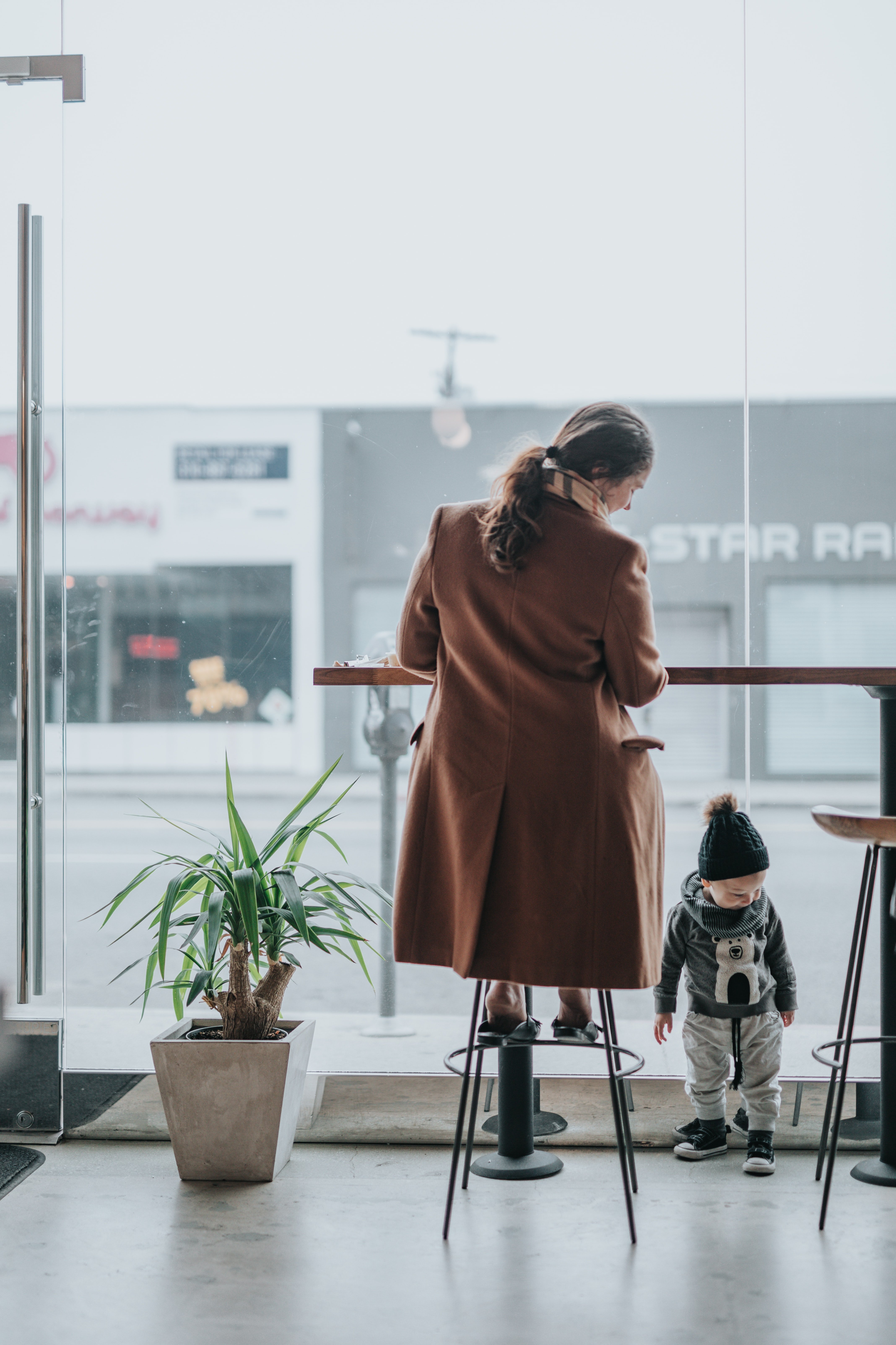 Regatta A woman sits at a high table in a café, while a small child plays nearby. A potted plant is visible in the foreground.