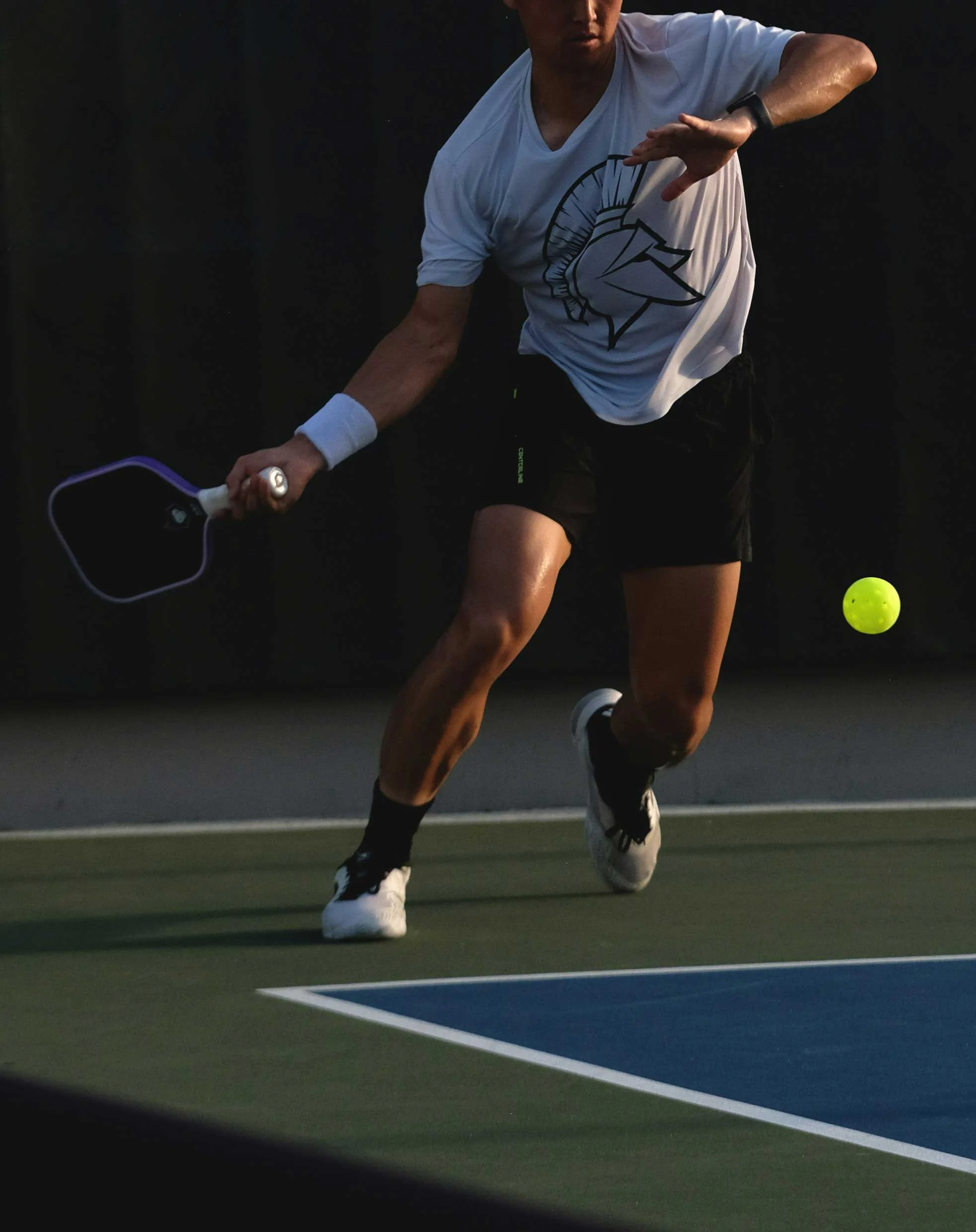 A player in a white shirt and black shorts is executing a forehand shot on a tennis court, with a green tennis ball in motion towards them. The background is dimly lit, highlighting the player’s action.