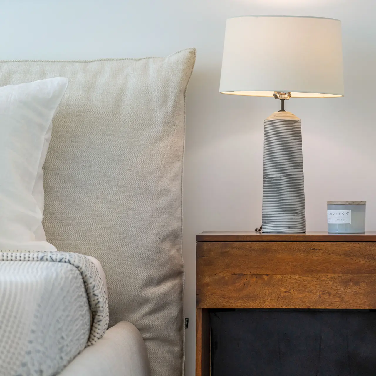 A stylish bedside table with a modern lamp and a candle, alongside a plush bed with a neutral-colored headboard and decorative pillows.