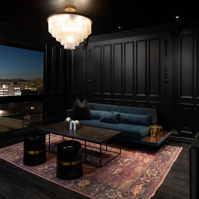 A modern living room with black walls, a plush teal sofa, stylish coffee tables, and a chandelier, overlooking a cityscape at dusk.