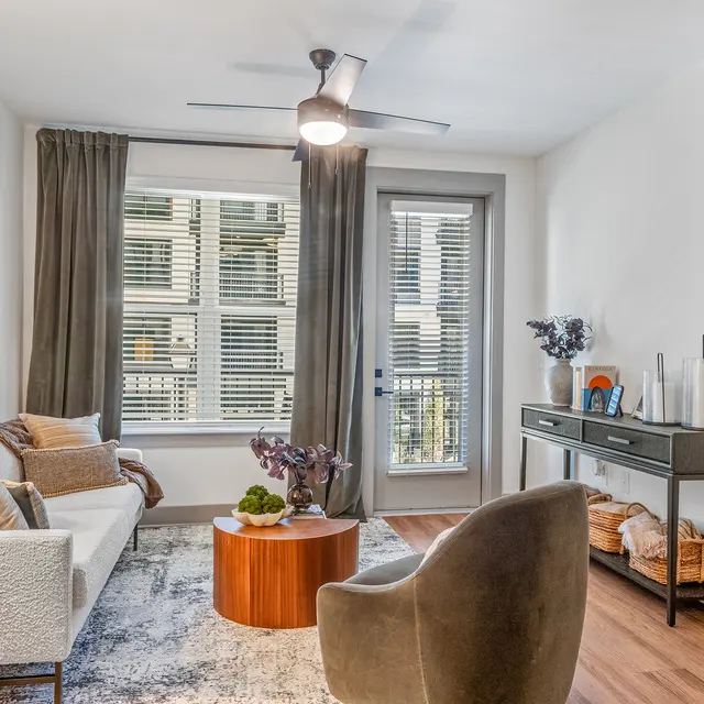 A modern living room featuring a white sofa with decorative pillows, a round wooden coffee table, and a stylish chair. The space is decorated with a large abstract painting, a floor lamp, and a console table displaying decorative items. Natural light enters through the large windows with curtains, and there is a door leading to a balcony.