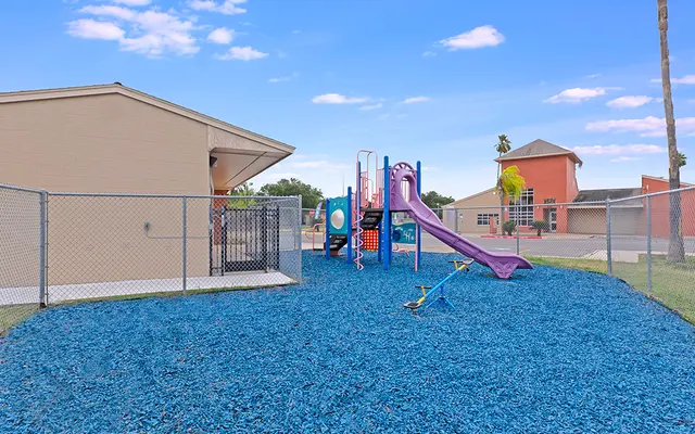 Children's Playground Area A fenced playground with a blue rubber surface, featuring a slide and climbing structure. Nearby buildings are visible under a clear blue sky.