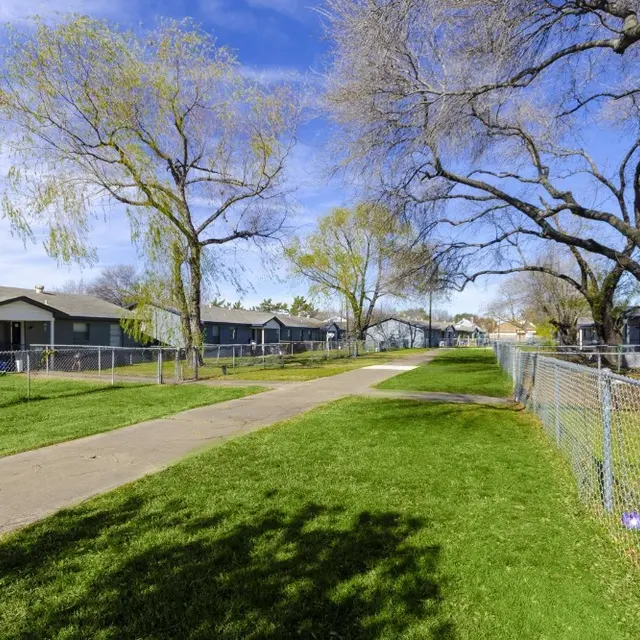 Park Pathway With Houses and Trees A paved pathway running through a grassy area bordered by houses and trees under a clear blue sky.
