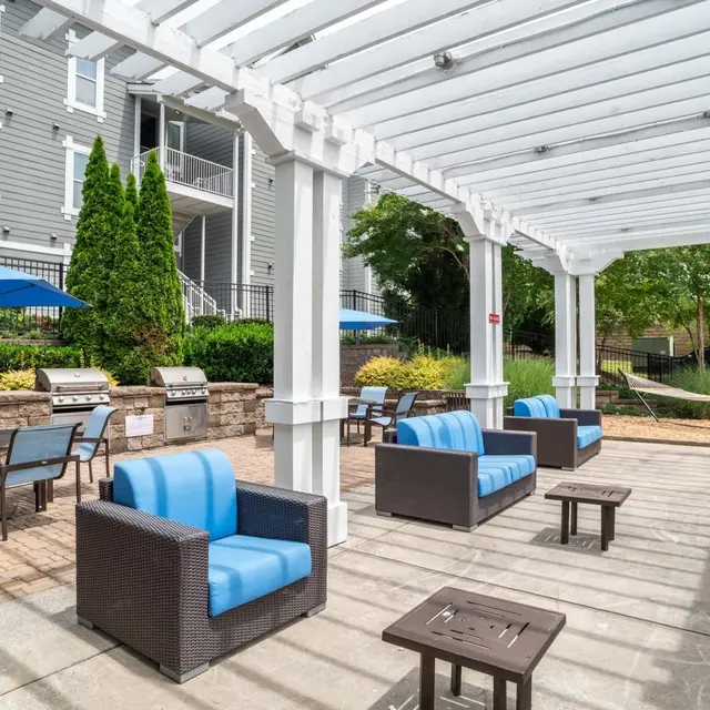 A modern outdoor lounge area featuring blue and brown wicker seating, small tables, and a grill station under a white pergola.