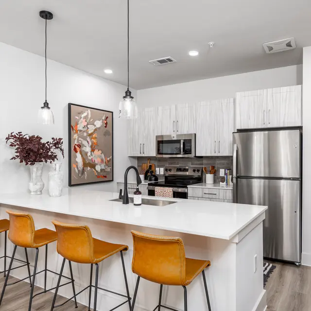 A modern kitchen featuring a white island with four brown bar stools, stainless steel appliances, and a beautiful artwork on the wall.