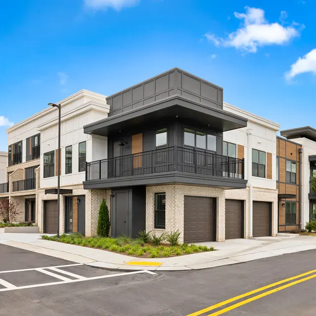 Modern Apartment Building Corner View A modern apartment building featuring a mix of materials including brick and dark panels, with balconies and garage doors, situated on a corner lot by a road.